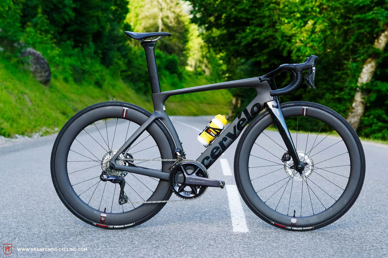 A sleek Cervelo road bike with a matte black frame on a quiet road surrounded by lush green trees.