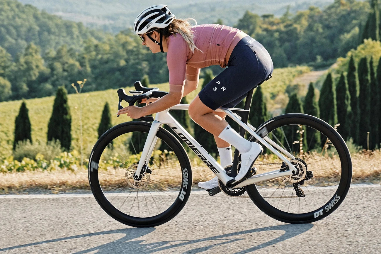 Cyclist wearing a helmet and pink jersey riding a white road bike on a scenic road.