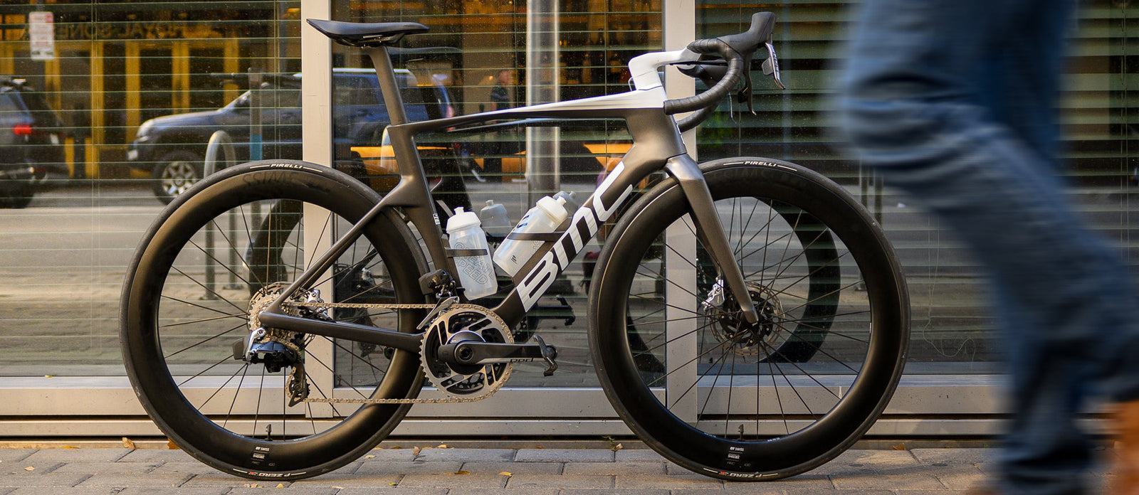 A sleek, high-performance road bike parked against a glass wall, with a person walking by in the foreground.