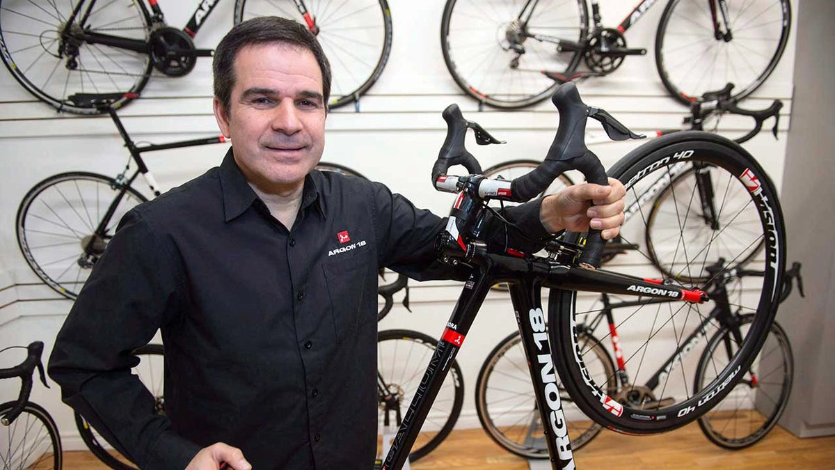 Man holding a road bike in a bicycle shop, with several bikes in the background.